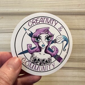 May include: A circular sticker with a cartoon illustration of a woman with purple hair and blue eyes. The sticker includes the words "CREATIVITY & COMMUNITY" and features a pencil and paintbrush. The woman has a tattoo of skulls.