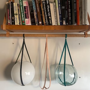 May include: A wooden shelf with books and hanging glass globe planters. The shelf is supported by leather straps. Two of the planters are white and one is teal, suspended by leather straps. The books are various sizes and colors.