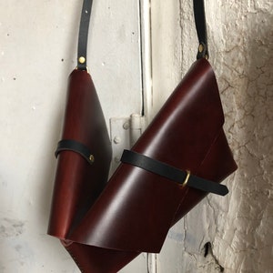 May include: Two dark burgundy leather bags with black straps and gold-colored hardware hang against a white wall. The bags are folded and secured with black leather straps. The leather has a glossy finish.