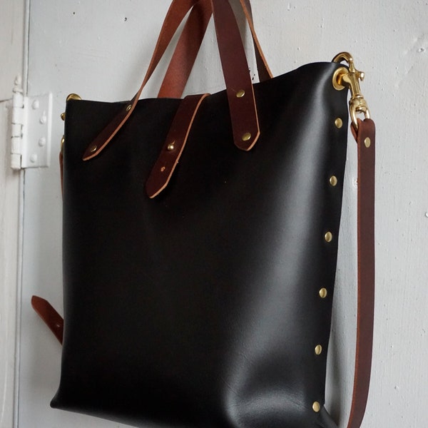 Può includere: Una borsa tote in pelle nera con cinturini in pelle marrone e dettagli in ottone. La borsa ha un manico superiore e una lunga tracolla.