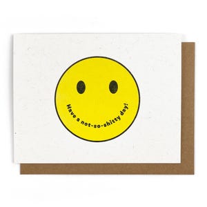 May include: A greeting card with a yellow smiley face and the text "Have a not-so-shitty day!" The card has a white front and a brown backing. The smiley face has black oval eyes and a black outline.