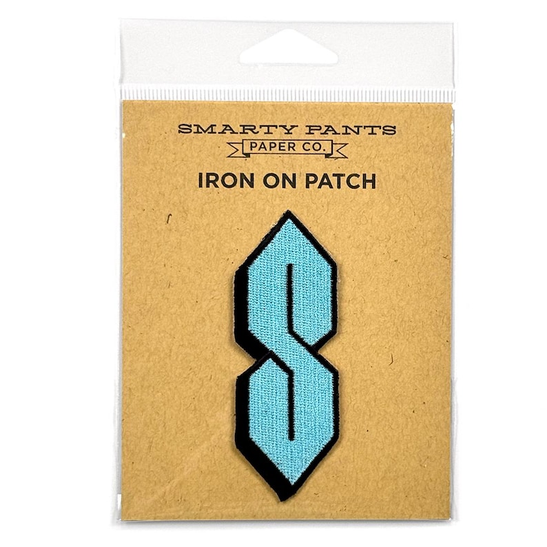 Op de afbeelding: Een lichtblauwe geborduurde strijkpatch in de vorm van een geometrische S, bevestigd op een beige kaart met het opschrift SMARTY PANTS PAPER CO. IRON ON PATCH. De patch heeft een scherp, hoekig ontwerp met een zwarte rand in een doorzichtige hoes.