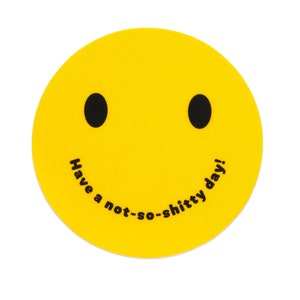 May include: A yellow, circular coaster with a smiley face design. The coaster features two black oval eyes and a curved black mouth. The phrase "Have a not-so-shitty day!" is printed in black along the bottom of the smiley face.