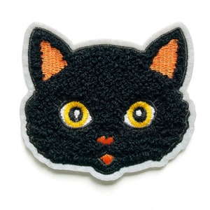 Peut inclure: Visage de chat en feutre noir avec des oreilles orange et des yeux jaunes. Le visage de chat est brodé sur un fond en feutre blanc.