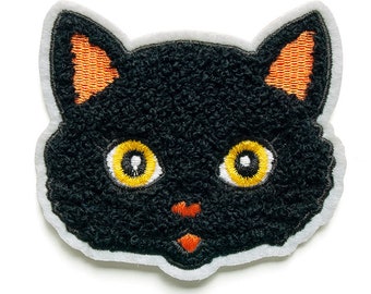 Può includere: Viso di gatto in feltro nero con orecchie arancioni e occhi gialli. Il viso del gatto è ricamato su uno sfondo in feltro bianco.