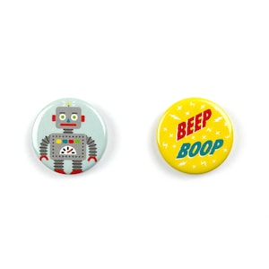 May include: Two round buttons. One button is blue with a cartoon robot on it. The robot is gray with red and yellow details. The other button is yellow with the words "BEEP BOOP" in red and blue letters.