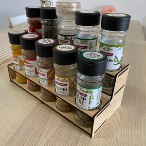May include: A wooden spice rack filled with glass jars of herbs and spices. The jars have black lids and labels with names such as "Cumin" and "Rosemary." The rack sits on a light wooden table, ideal for kitchen storage.