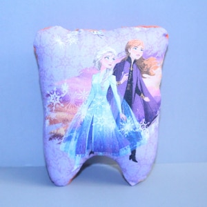 Könnte beinhalten: Ein zahnförmiges Kissen mit einer farbenfrohen Illustration von zwei Charakteren aus dem Film Frozen. Das Kissen hat einen lila Hintergrund mit Schneeflockenmustern und einen hellorangen Rand. Die Charaktere tragen blaue und violette Outfits.