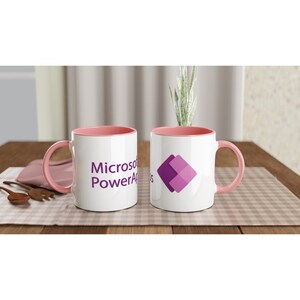 May include: Two white ceramic mugs with pink rims. One mug has the text "Microsoft PowerApps" printed on it. The other mug has a purple logo of two overlapping squares.