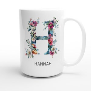 May include: White ceramic mug with a floral design in shades of blue, pink, and yellow. The letter "H" is formed by the flowers. The name "HANNAH" is printed below the floral design.