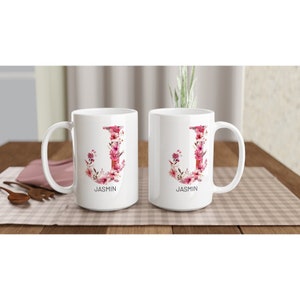 May include: Two white ceramic mugs with a floral design in shades of pink and white. The letter "J" is formed by the flowers and the name "JASMINE" is printed below each mug.