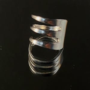 May include: A silver-colored ring crafted from repurposed silverware. The ring features a unique design with multiple curved bands and a rectangular top, reflecting light on a dark surface. A creative and artistic jewelry piece.