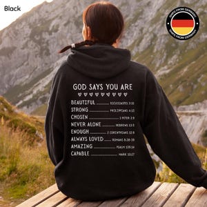 Könnte beinhalten: Schwarzer Kapuzenpullover mit weißem Text: "GOD SAYS YOU ARE" und einer Liste von Affirmationen und Bibelversen. Der Kapuzenpullover wird von einer Person mit dunklen Haaren getragen, die sich im Freien aufhält. Das Bild enthält ein "Versand aus Deutschland"-Logo.