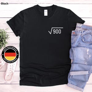 Op de afbeelding: Een zwart t-shirt met korte mouwen en een witte afbeelding op de linkerborst. De afbeelding toont een vierkantswortelsymbool gevolgd door het getal 900. Het t-shirt heeft een ronde hals en een standaard pasvorm op een plat oppervlak.