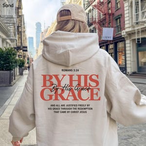 May include: A sand-colored hoodie with the text "ROMANS 3:24" and "BY HIS GRACE" in red. Below the text is the phrase "AND ALL ARE JUSTIFIED FREELY BY HIS GRACE THROUGH THE REDEMPTION THAT CAME BY CHRIST JESUS". A beige baseball cap is also visible.
