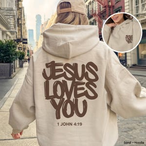 Può includere: Una felpa con cappuccio color sabbia con il testo marrone scuro JESUS LOVES YOU e il riferimento 1 JOHN 4:19 stampati sul retro. Una versione più piccola dello stesso testo è stampata sulla parte superiore sinistra del petto della felpa.