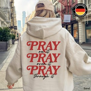 Op de afbeelding: Een zandkleurige hoodie met de rode tekst "PRAY on it, PRAY over it, PRAY through it". De hoodie heeft een capuchon en wordt door een persoon gedragen. Een beige baseballpet is ook zichtbaar.