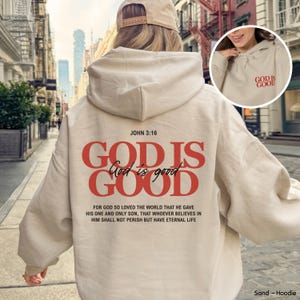 Può includere: Una felpa con cappuccio color sabbia con testo rosso sul retro che recita "JOHN 3:16 GOD IS GOOD" in grassetto, con "God is good" in corsivo e il versetto biblico completo sottostante. Una versione più piccola è presente sul davanti.