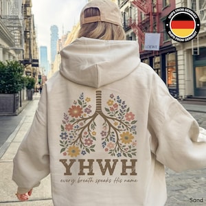 Może przedstawiać: Bluza z kapturem w kolorze piaskowym z kwiatowym wzorem płuc na plecach. Wzór przedstawia kolorowe kwiaty i napis "YWHW every breath speaks His name". Bluza ma kaptur ze sznurkiem i kieszeń typu kangur.