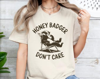 May include: A beige t-shirt with a brown graphic of a honey badger wearing sunglasses and relaxing in a lounge chair. The text "Honey Badger Don't Care" is printed below the graphic.