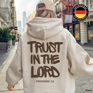 Könnte beinhalten: Sandfarbener Kapuzenpullover mit dem Text "TRUST IN THE LORD" und "PROVERBS 3:5" in Braun aufgedruckt. Der Hoodie hat eine Kapuze und lange Ärmel. Eine Baseballkappe wird getragen. Der Hintergrund zeigt eine Stadtstraße.