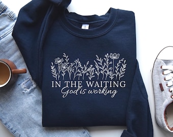 May include: A navy blue crewneck sweatshirt featuring a white line art graphic of various wildflowers across the chest. Below the floral design, the text reads "IN THE WAITING" in a bold sans-serif font, followed by "God is working" in a cursive script.