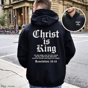 Op de afbeelding: Een zwarte hoodie met witte tekst op de achterkant die Christ is King leest, gevolgd door het vers Revelation 19:16 en de volledige bijbeltekst. Het borstgedeelte toont ook de witte tekst Christ is King.