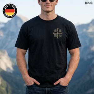 May include: A black short-sleeved t-shirt featuring a gold-colored cross graphic on the left chest. The design includes the text IC XC NI KA arranged around the cross. The shirt has a crew neckline and a standard fit, presented in a solid black color.