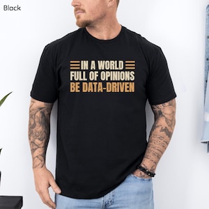 May include: Black t-shirt with the text "IN A WORLD FULL OF OPINIONS BE DATA-DRIVEN" in a tan and brown font. The shirt is a crew neck and short-sleeved.