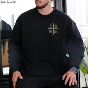 Könnte beinhalten: Ein schwarzes Sweatshirt mit Rundhalsausschnitt und einem beigen Kreuz-Motiv auf der linken Brust. Das Kreuz-Design enthält den Text IC XC NI KA, der in den vier Quadranten angeordnet ist. Das Sweatshirt hat lange Ärmel sowie gerippte Bündchen.