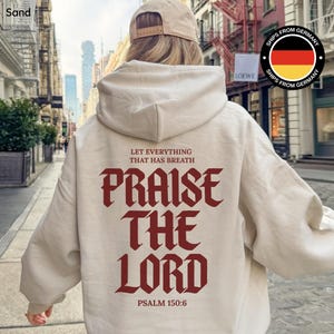 May include: Sand-colored hoodie with a hood and long sleeves. The back of the hoodie features the text "LET EVERYTHING THAT HAS BREATH PRAISE THE LORD" in a burgundy gothic font, with "PSALM 150:6" below. A beige baseball cap is worn.