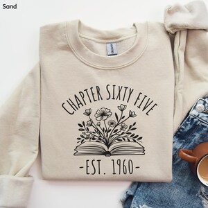 May include: Sand-colored sweatshirt with the text "CHAPTER SIXTY FIVE" above an open book with floral accents. Below the book is "-EST. 1960-". The sweatshirt is folded, with a portion of a denim garment visible.