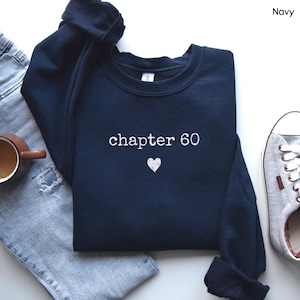 May include: A navy blue crewneck sweatshirt featuring white embroidered text that reads chapter 60 with a small white heart icon centered below the text. The sweatshirt is displayed alongside light blue denim jeans and a grey sneaker.