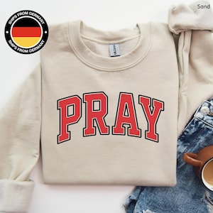 Op de afbeelding: Een zandkleurige sweatshirt met het woord "PRAY" in rode blokletters met een zwarte omlijning. De sweatshirt heeft een ronde hals en lange mouwen. De afbeelding toont ook een spijkerbroek en een koffiekopje.