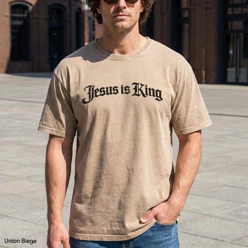 May include: A beige t-shirt with the text "Jesus is King" in a black, stylized font. The shirt is a neutral color and has short sleeves. The image shows the shirt on a person.