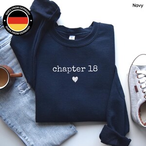 Op de afbeelding: Marineblauw sweatshirt met de tekst "chapter 18" en een hartje. Het sweatshirt wordt gecombineerd met een blauwe jeans en grijze sneakers. Een koffiemok is links zichtbaar.