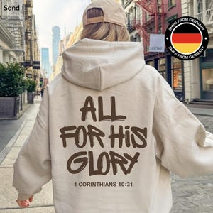 May include: A sand-colored hoodie with the text "ALL FOR HIS GLORY" in brown lettering on the back. The text "1 Corinthians 10:31" is below the main text. The person is wearing a tan baseball cap.