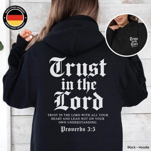 Könnte beinhalten: Ein schwarzer Kapuzenpullover mit weißem Text auf der Rückseite, der Trust in the Lord lautet, gefolgt vom vollständigen Vers aus Sprüche 3,5. Ein kleinerer kreisförmiger Einsatz zeigt die Vorderseite desselben schwarzen Hoodies mit dem Text auf der Brust.