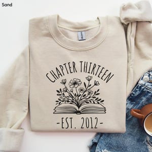 May include: Sand-colored sweatshirt with the text "CHAPTER THIRTEEN" above an open book illustration with flowers and the text "-EST. 2012-". The sweatshirt is folded and placed on a denim surface.