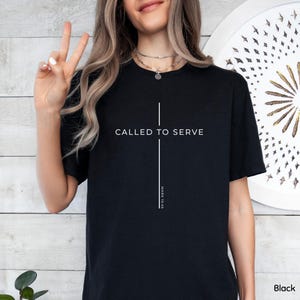 May include: Black t-shirt with the text "CALLED TO SERVE" and "MARK 10:45" in white. The design features a vertical line intersecting a horizontal line, forming a cross. The shirt is a solid black color.