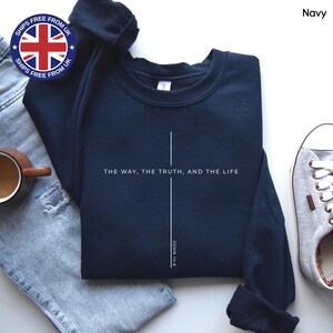 May include: Navy blue sweatshirt featuring the text "THE WAY, THE TRUTH, AND THE LIFE" and "JOHN 14:6" in white. The design includes a simple cross. The image also shows a pair of jeans, a coffee mug, and a sneaker.
