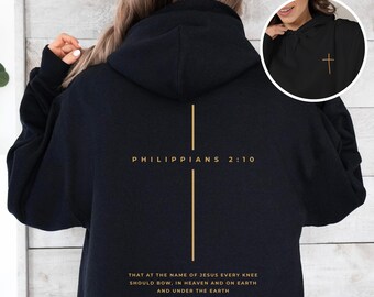 May include: A black hoodie featuring a gold cross graphic on the back with the text Philippians 2:10 and the verse That at the name of Jesus every knee should bow, in heaven and on earth and under the earth. A small gold cross is on the front.
