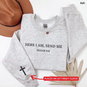 May include: A light gray sweatshirt with the text "HERE I AM, SEND ME ISAIAH 6:8" in black. A black cross is on the left sleeve. A gold necklace with a pendant is on the sweatshirt. A brown hat is in the background.