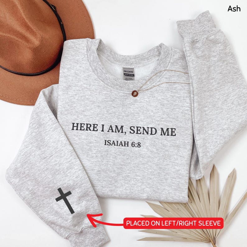 May include: A light gray sweatshirt with the text "HERE I AM, SEND ME ISAIAH 6:8" in black. A black cross is on the left sleeve. A gold necklace with a pendant is on the sweatshirt. A brown hat is in the background.