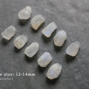 Puede incluir: Un conjunto de peque&ntilde;as piedras transl&uacute;cidas y p&aacute;lidas, posiblemente piedras lunares, sobre una superficie gris. Las piedras tienen forma irregular y parecen estar talladas toscamente. El texto en la parte inferior dice "stone size: 12-14mm" y "crystalsofart".