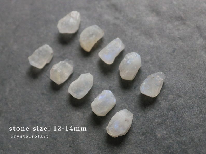 Puede incluir: Un conjunto de peque&ntilde;as piedras transl&uacute;cidas y p&aacute;lidas, posiblemente piedras lunares, sobre una superficie gris. Las piedras tienen forma irregular y parecen estar talladas toscamente. El texto en la parte inferior dice "stone size: 12-14mm" y "crystalsofart".