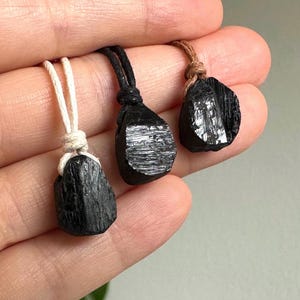 May include: Three black tourmaline pendants with natural, rough-hewn surfaces. Each stone is suspended from a cord: one white, one black, and one brown. The pendants are displayed against a neutral background.