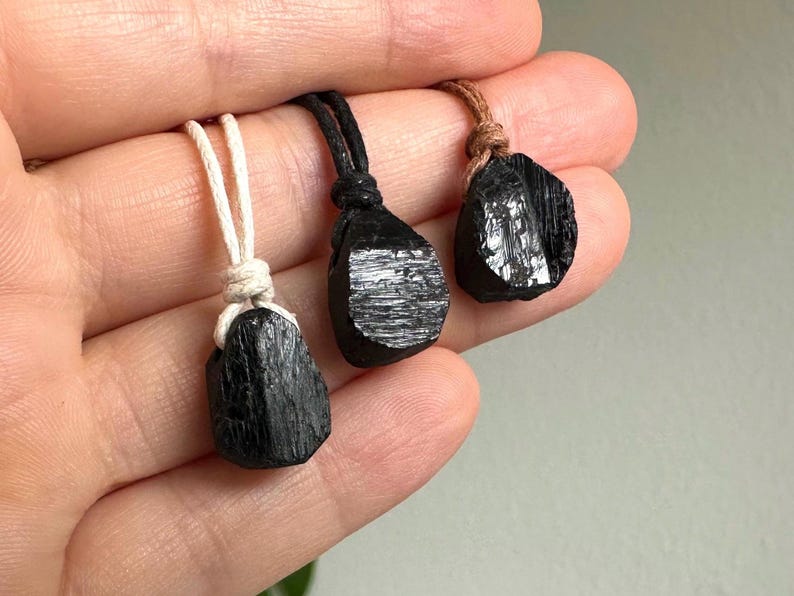 May include: Three black tourmaline pendants with natural, rough-hewn surfaces. Each stone is suspended from a cord: one white, one black, and one brown. The pendants are displayed against a neutral background.