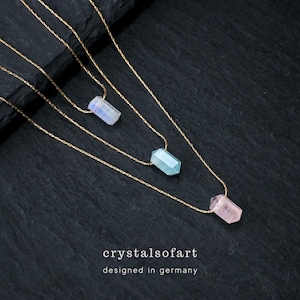 May include: Three gold chain necklaces, each with a different gemstone pendant: a white, a light blue, and a pink crystal. The necklaces are displayed on a dark, textured surface. The text "crystalsofart" and "designed in germany" are at the bottom.