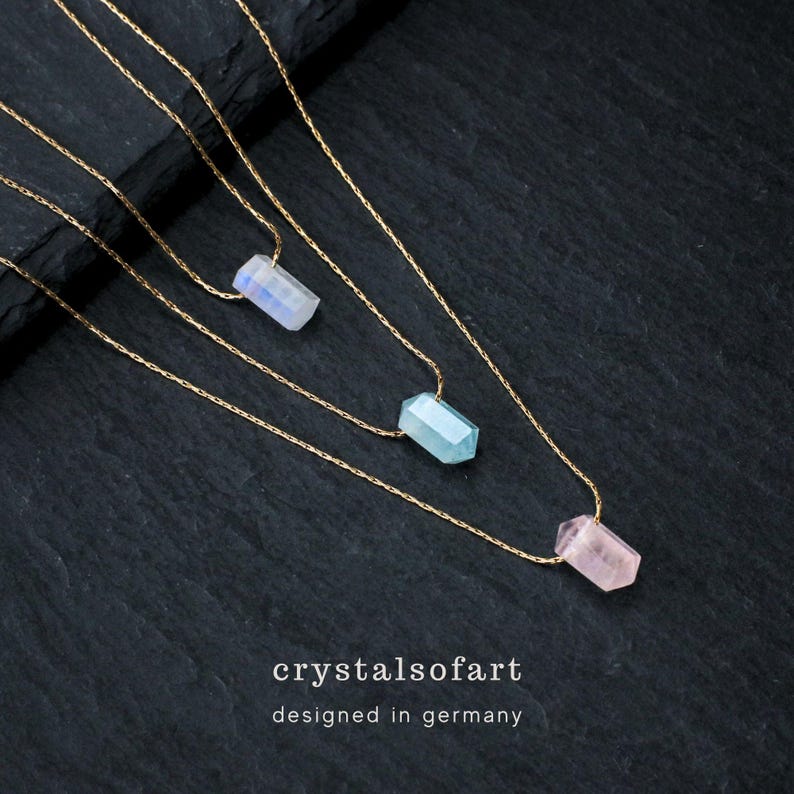 May include: Three gold chain necklaces, each with a different gemstone pendant: a white, a light blue, and a pink crystal. The necklaces are displayed on a dark, textured surface. The text "crystalsofart" and "designed in germany" are at the bottom.